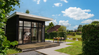 Moderne glamping-lodge met grote ramen, terras en buitentafel bij een vijver onder een zonnige hemel.