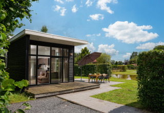 Cabaña glamping moderna con grandes ventanales, terraza y comedor exterior junto a un estanque bajo cielo azul.
