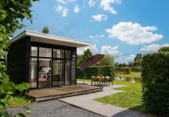 Moderne glamping-hytte med store vinduer, terrasse og udendørs spisested ved dam under blå himmel.