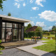 Cabaña glamping moderna con grandes ventanales, terraza y comedor exterior junto a un estanque bajo cielo azul.