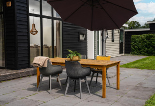 Buiten eethoek bij glamping met houten tafel, stoelen, grote parasol en zicht op modern vakantiehuisje.