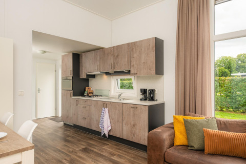 Cucina moderna in alloggio glamping con mobili in legno, divano e ampia finestra sul verde.