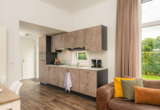 Modern glamping accommodation kitchen with wood cabinets, couch, and large window overlooking greenery.