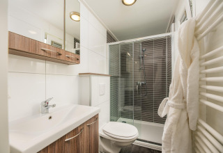 Moderne badkamer bij een glampingverblijf met douche, wastafel, toilet, houtkasten en witte badjas.