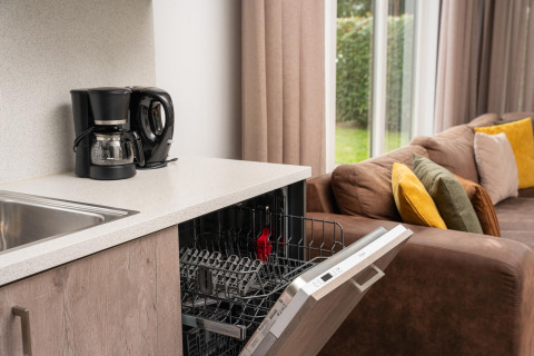 Moderne glamping accommodatie met koffiezetapparaat, open vaatwasser en bank met kleurrijke kussens bij het raam.