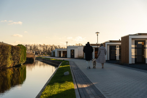 Twee mensen en een hond wandelen langs moderne lodges bij een kanaal op een zonnige, heldere dag.