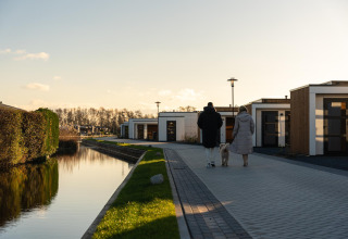 Twee mensen en een hond wandelen langs moderne lodges bij een kanaal op een zonnige, heldere dag.