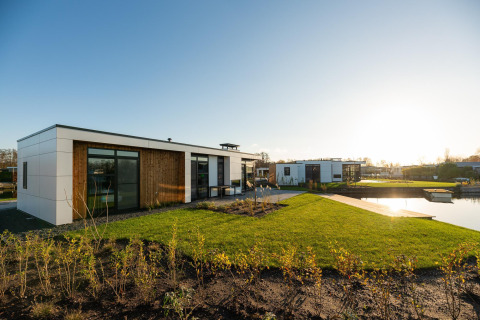 Moderne Villa Nautica-lodge met ruime glaspartijen, grasveld en vijver in de avondzon.