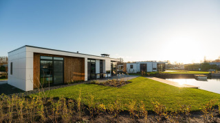 Moderna cabaña Villa Nautica con grandes ventanales y césped junto a un estanque al atardecer.