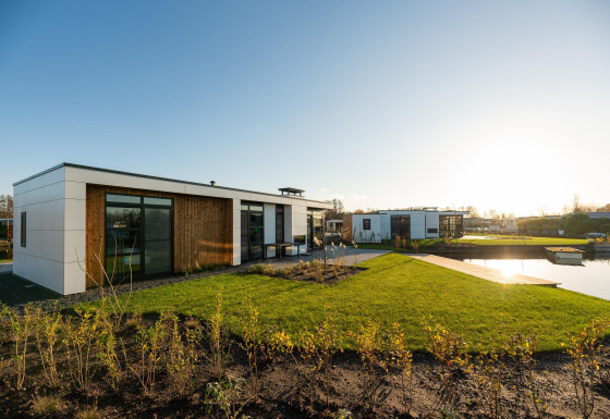 Moderne Villa Nautica-lodge met ruime glaspartijen, grasveld en vijver in de avondzon.