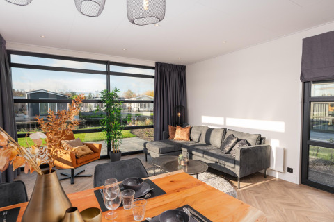 Lyst opholdsområde i Villa Nautica-lodgen med store vinduer, moderne sofa og spisebord udsigt til natur.