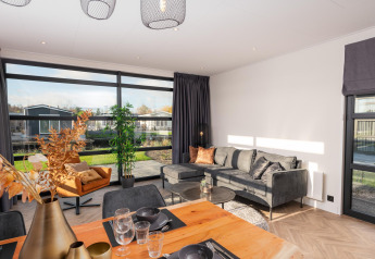 Lyst opholdsområde i Villa Nautica-lodgen med store vinduer, moderne sofa og spisebord udsigt til natur.