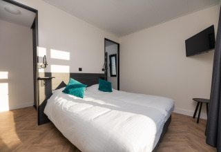 Lichte slaapkamer in Villa Nautica lodge met tweepersoonsbed, twee groene kussens en wand-tv.