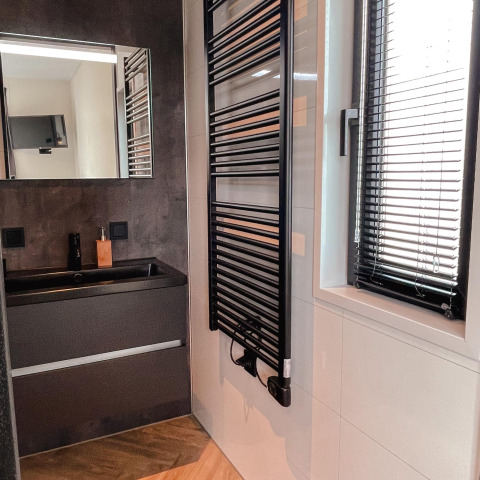 Baño moderno con lavabo negro, espejo de pared, toallero calefaccionado y ventana con persianas negras.