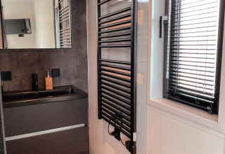 Baño moderno con lavabo negro, espejo de pared, toallero calefaccionado y ventana con persianas negras.