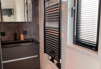 Baño moderno con lavabo negro, espejo de pared, toallero calefaccionado y ventana con persianas negras.