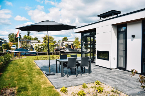 Casa moderna con terraza, muebles de jardín y sombrilla, ubicada tranquilamente junto al agua.