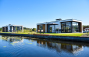 Lodges modernos de Villa Nautica con grandes ventanales y terrazas frente a un tranquilo canal bajo cielo azul.