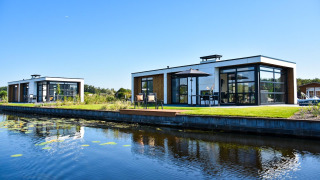 Lodges modernes Villa Nautica avec grandes baies vitrées et terrasse au bord de l’eau sous un ciel bleu clair.