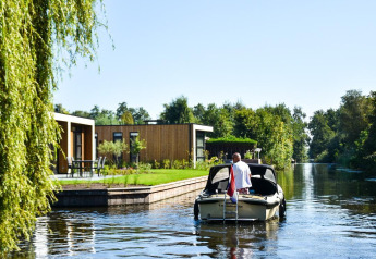 En mand sejler i en lille båd ved Villa Nautica-hytten, med moderne villaer og træer i baggrunden.