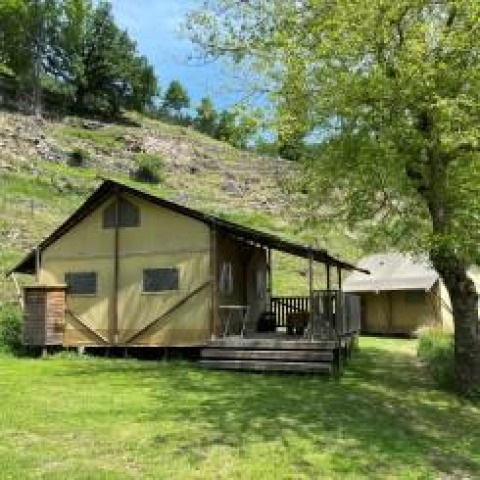 Foto di una tenda safari con veranda in legno su un prato verde, circondata da alberi e una collina.