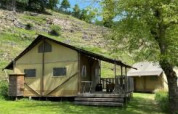 Foto di una tenda safari con veranda in legno su un prato verde, circondata da alberi e una collina.