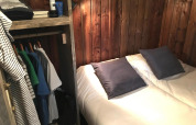 Hyggeligt glampingværelse med træpaneler, dobbeltseng, to puder og åben garderobe med tøj.