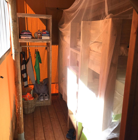 Alojamiento glamping con literas de madera, mosquitera y estante abierto con ropa y cesta en la tienda.