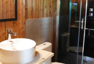 Baño moderno en alojamiento glamping con paredes de madera, lavabo redondo, inodoro y ducha acristalada.