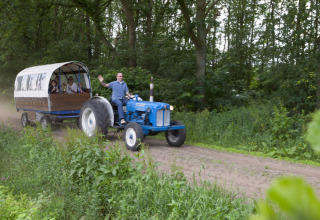 Man bestuurt blauwe tractor met huifkar vol mensen op bosweg van Feather Down De Lange Weide, Drenthe.
