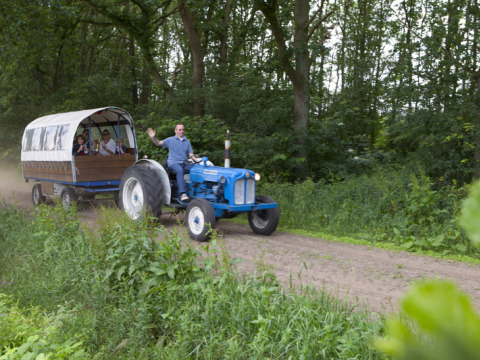Man rijdt op blauwe tractor met huifkar vol mensen over bospad bij Feather Down De Lange Weide, Drenthe.