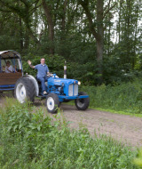 Hombre conduce tractor azul con remolque cubierto lleno de personas en Feather Down De Lange Weide, Drenthe.