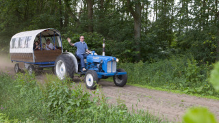 Hombre conduce tractor azul con remolque cubierto lleno de personas en Feather Down De Lange Weide, Drenthe.