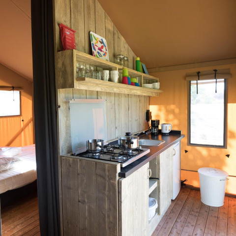 Pequeña cocina en una tienda safari con estufa de gas, estantes abiertos, utensilios y vista al dormitorio.
