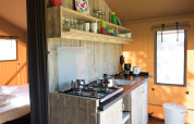 Pequeña cocina en una tienda safari con estufa de gas, estantes abiertos, utensilios y vista al dormitorio.