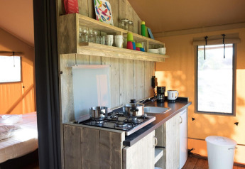 Pequeña cocina en una tienda safari con estufa de gas, estantes abiertos, utensilios y vista al dormitorio.