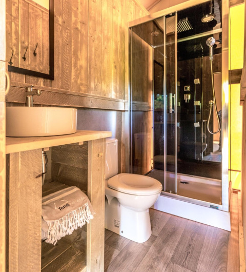 Baño luminoso en una tienda safari con paneles de madera, ducha moderna, inodoro y lavabo de cerámica.