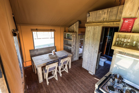 Interior de tienda safari con mesa de madera, sillas, literas, cocina básica y baño moderno.