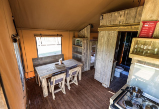 Interior de tienda safari con mesa de madera, sillas, literas, cocina básica y baño moderno.