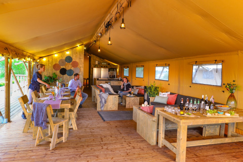 Intérieur d'une grande tente safari avec meubles en bois, coin salon et personnes prenant un repas ensemble.