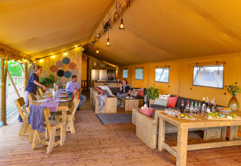 Interior of a spacious safari tent with wooden furniture, lounge area, and people dining and relaxing.