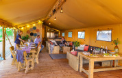 Intérieur d'une grande tente safari avec meubles en bois, coin salon et personnes prenant un repas ensemble.