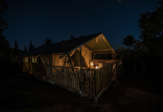 Tente safari éclairée sur une terrasse en bois, sous un ciel étoilé nocturne et entourée d’arbres.