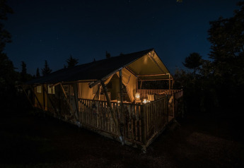 Tenda safari illuminata con veranda in legno di notte, immersa tra alberi e cielo stellato.