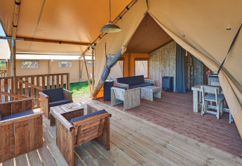 Interno di una tenda safari con mobili in legno, zona salotto, tavolo da pranzo e veranda aperta.