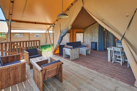 Binnenzicht van een safaritent met houten meubels, zithoek, eettafel en open veranda in een groene omgeving.