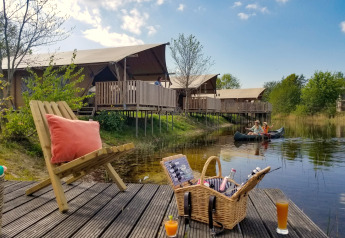 Safari tent glamping by a lake, with canoe, picnic basket, and drinks on a sunlit wooden deck.