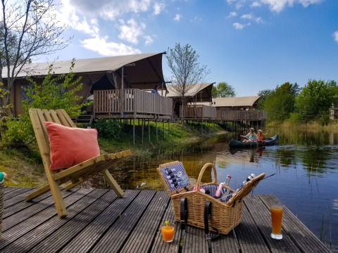 Campamento con tiendas safari junto al lago, canoa, cesta de picnic y bebidas en terraza de madera.