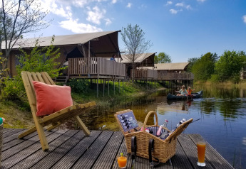 Campamento con tiendas safari junto al lago, canoa, cesta de picnic y bebidas en terraza de madera.