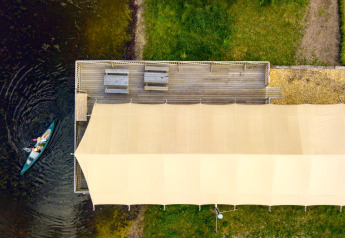Aerial view of a safari tent by a lake, a canoe approaching the dock, and benches on a wooden deck.